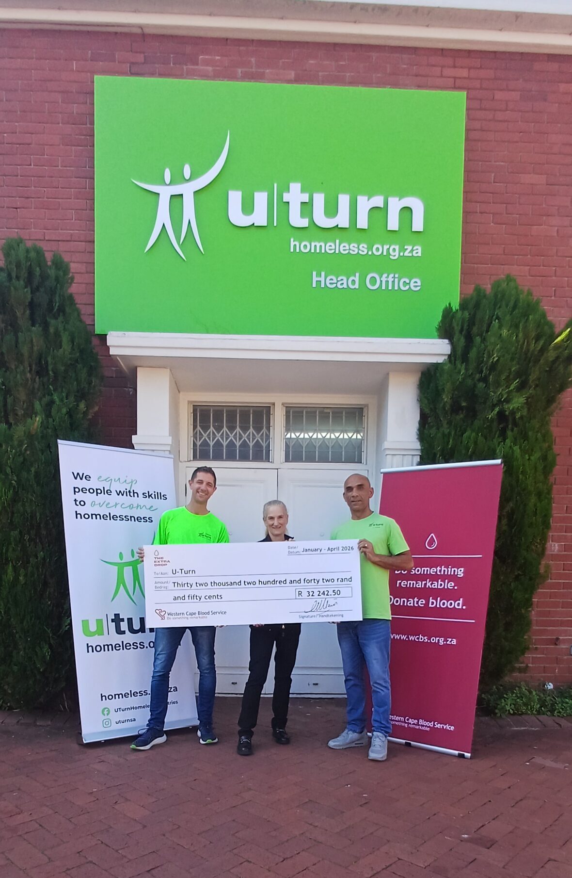 Three people in neon green shirts hold a large ceremonial cheque outside a brick building with a bright green U-turn sign above them.