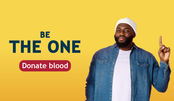 Smiling man in a white beanie and denim shirt points up beside bold text 'Be The One' and a red 'Donate blood' badge on a yellow gradient background.